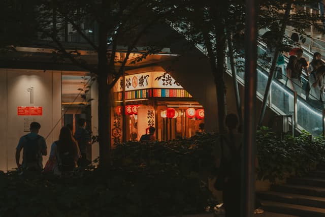 A nighttime street scene with a warmly lit restaurant featuring red lanterns, people walking nearby, and an illuminated staircase on the right