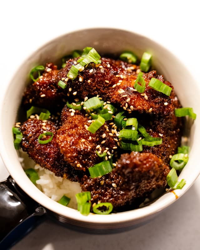 A bowl of crispy coated food topped with green scallions and sesame seeds, served over a bed of rice