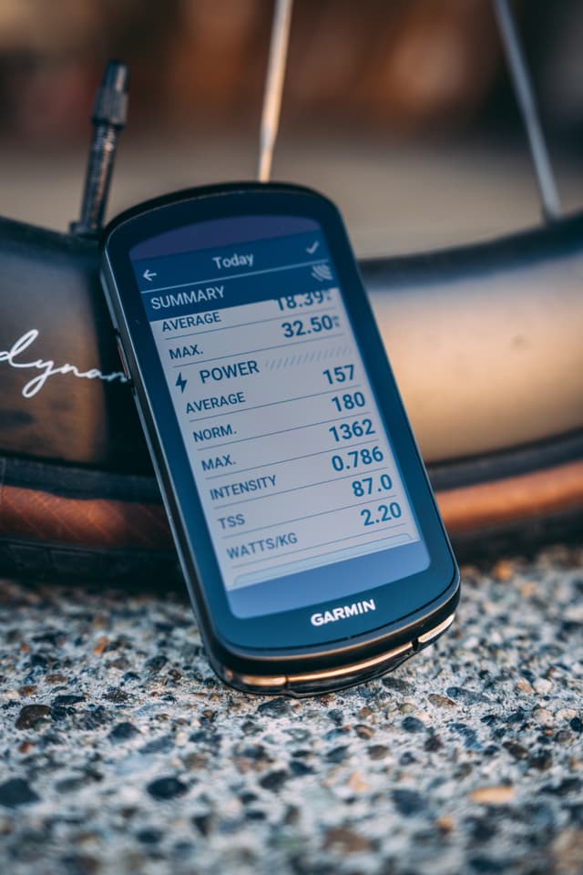 A Garmin cycling computer displays summary statistics, including speed, power, and intensity, resting against a bicycle wheel on a gravel surface