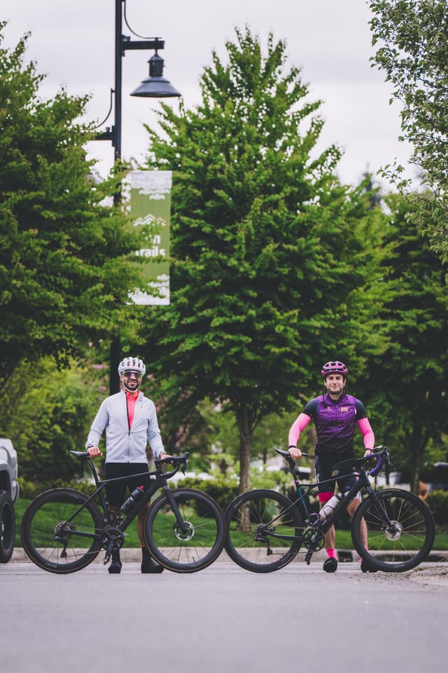 Two cyclists wearing helmets and cycling gear stand with their bikes on a paved path, surrounded by lush green trees