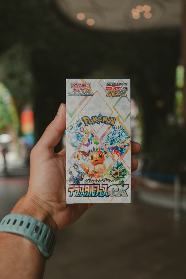 A hand holding a Pokémon card box with colorful artwork and Japanese text, set against a blurred outdoor background