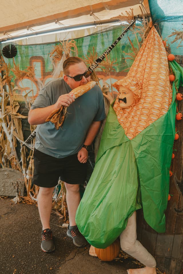 A man playfully pretends to eat corn on the cob while standing next to someone dressed in a corn costume. They are both in a festive, rustic setting with corn stalks and autumn-themed decorations