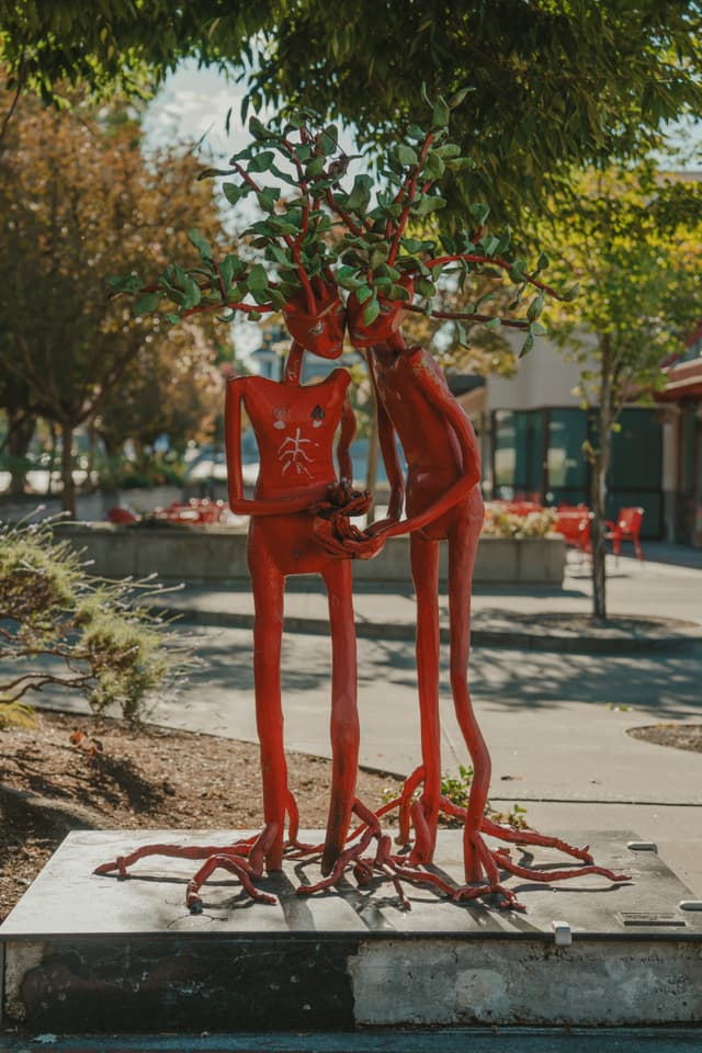 Two red, tree-like sculptures with human forms stand intertwined, featuring branches and leaves as heads, set in an outdoor urban environment