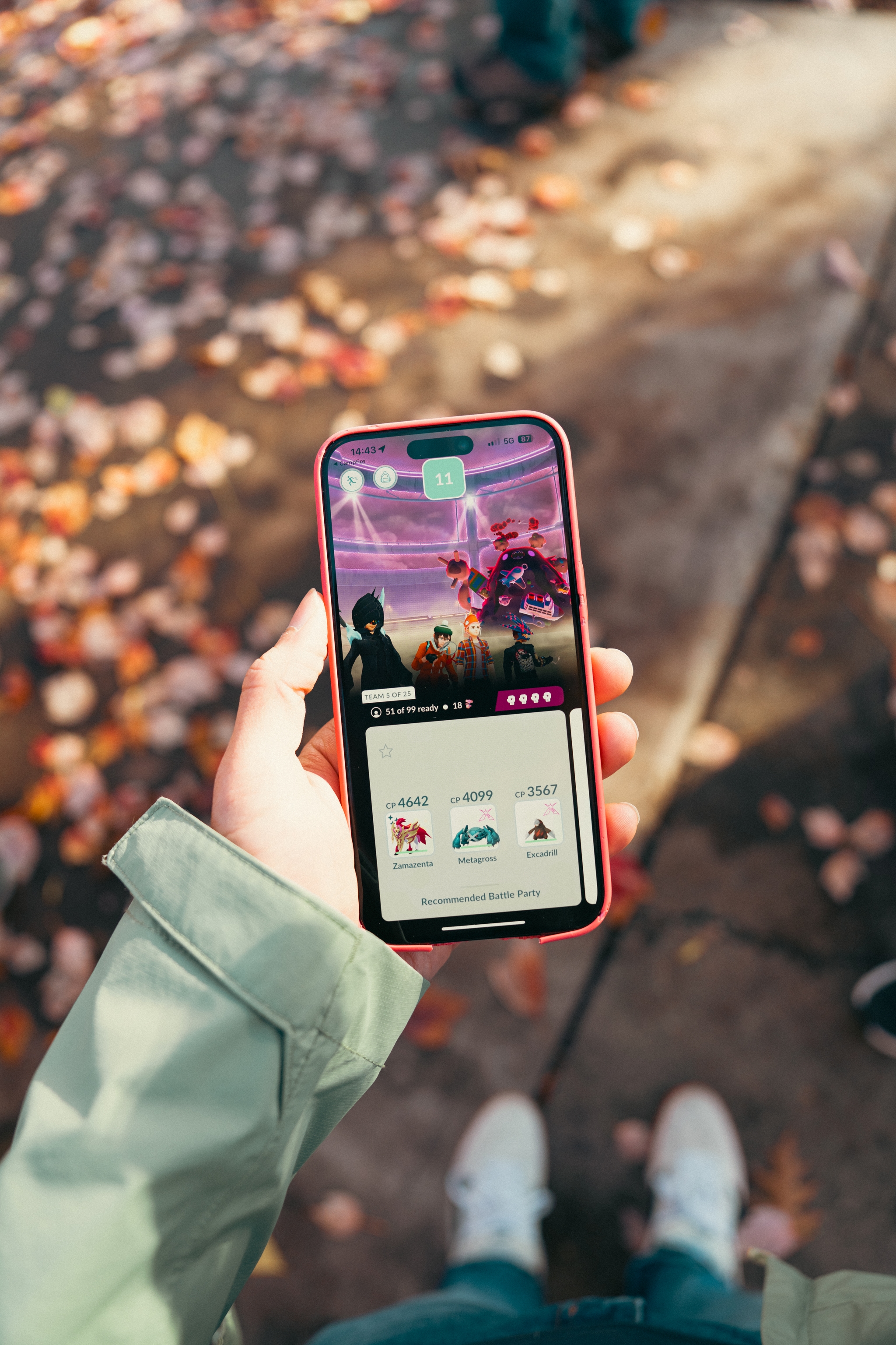 Person holding a smartphone displaying a Pokémon game, with fallen autumn leaves on the ground