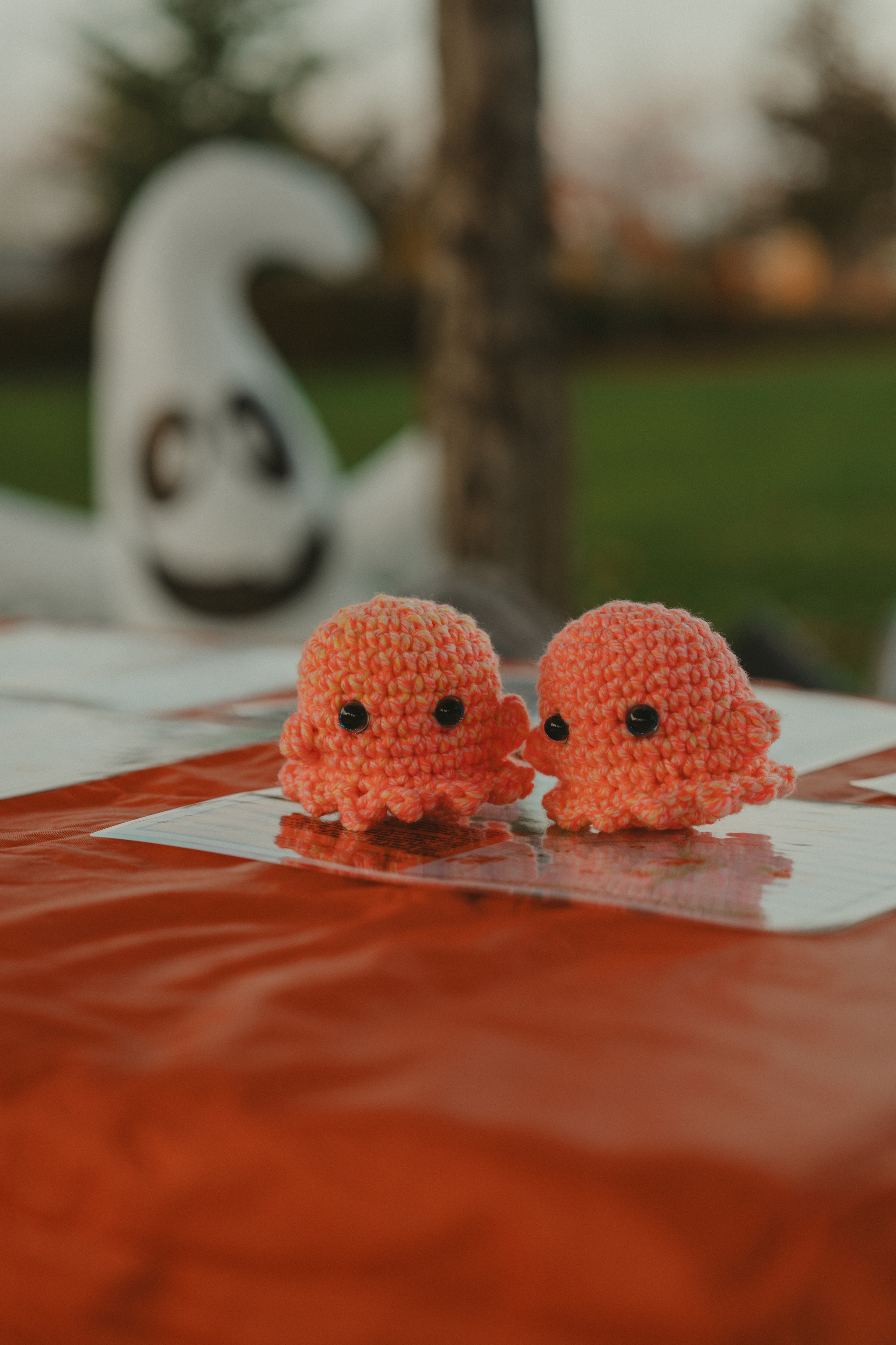 Two small crochet ghosts with large eyes sitting on a red surface outdoors with a ghost decoration in the background