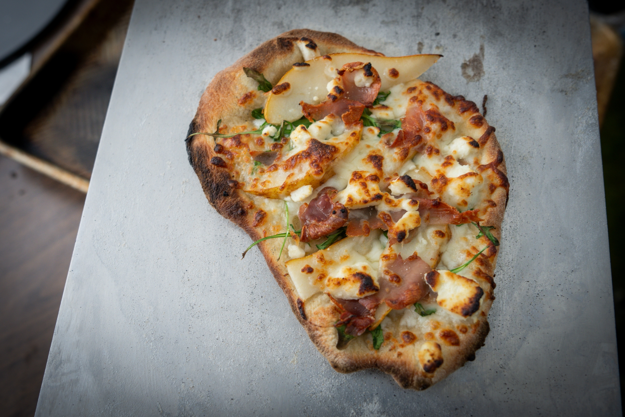 A rustic, irregularly shaped pizza topped with cheese, prosciutto, and herbs on a metal surface