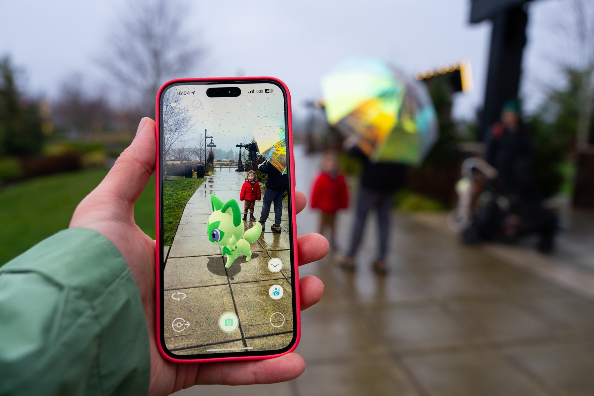 A person holding a smartphone with an augmented reality game displaying a green creature on the screen In the background, people are walking on a wet sidewalk, one holding a colorful umbrella